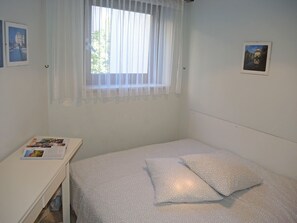 Ein kleines Zimmer mit einem Einzelbett, einem Fenster mit durchsichtigen Vorhängen, einem Nachttisch mit einer Zeitschrift und einem Wandbild.