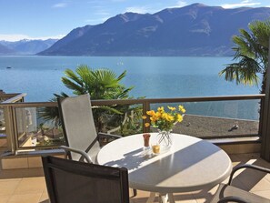 Mesa redonda com um vaso de flores amarelas, uma cadeira e vista para um lago calmo com montanhas ao fundo.