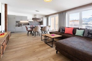 A modern living area with a sofa, a coffee table, and a dining table with chairs. There is a kitchen in the background, and large windows provide a view of the outdoors. - Holiday apartment in Riezlern with Wi-Fi (Riezlern)