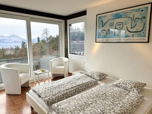 Ein Schlafzimmer mit zwei Betten, einem Sessel, einem kleinen Tisch und einem großen Fenster mit Blick auf Bäume und Berge.