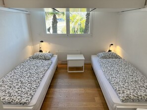 A small, compact room with two single beds, a small white table, and a window with a view of greenery. - Residenza Delphin by Interhome (Brissago)
