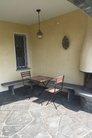 An outdoor patio with a stone floor, a wooden table, two chairs, a bench, a wall-mounted light fixture, and a decorative wall piece. - La Civetta by Interhome (Brissago)