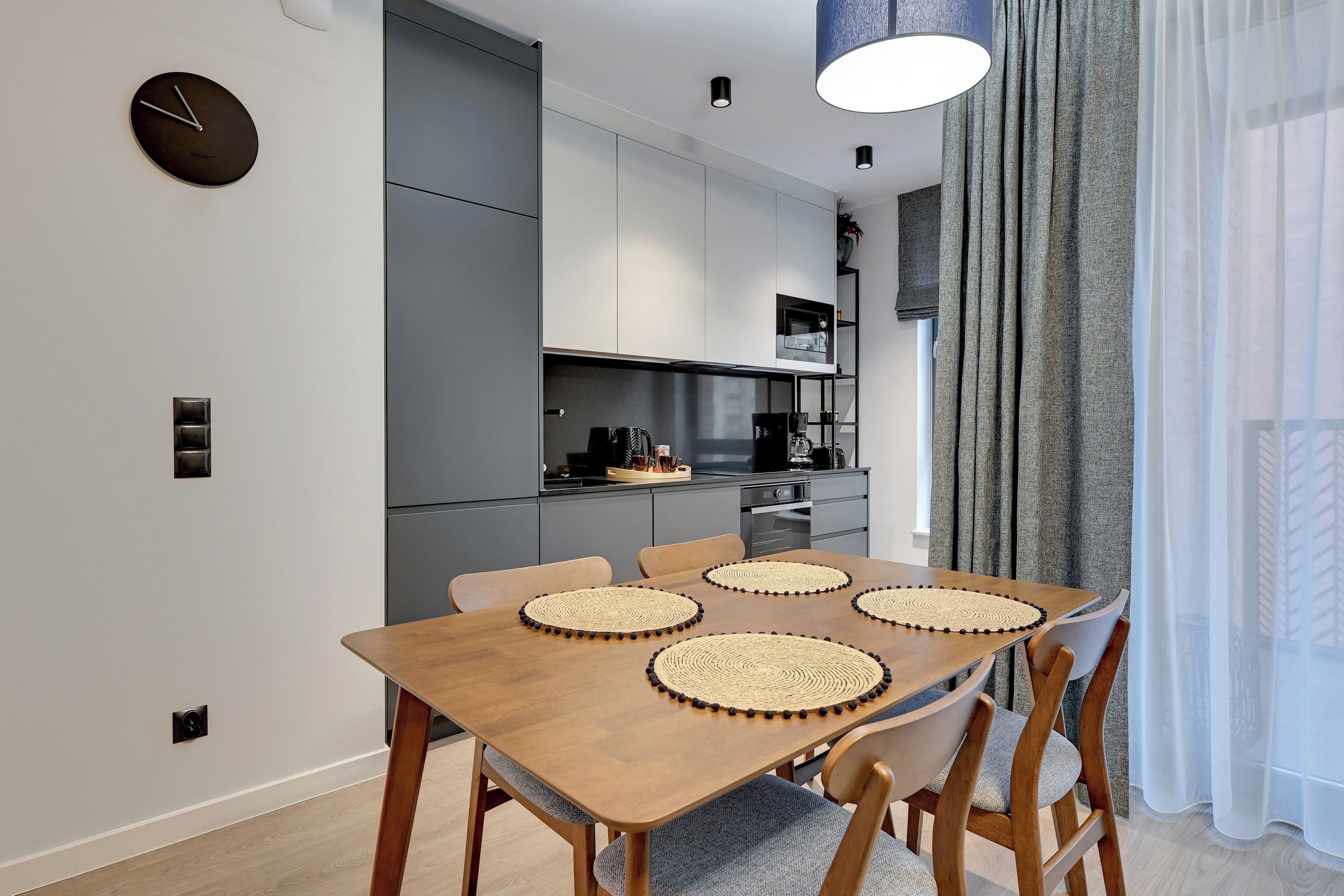 Une cuisine moderne avec des meubles de couleur grise, une table à manger en bois et des sets de table en fibres tressées, ainsi qu’une horloge noire accrochée au mur.