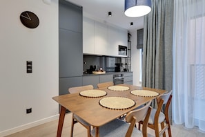 Een moderne keuken met grijze kastjes, een houten eettafel met geweven placemats en een zwarte klok aan de muur.