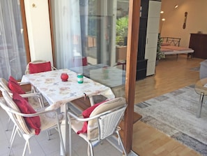 Ein Essbereich mit Glastisch und Korbstühlen, eine Küche im Hintergrund und ein kleiner Balkon mit einer Couch.