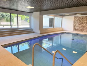 Ein moderner, überdachter Schwimmbereich mit Glaswänden, Holzdecke und Blick ins Grüne.