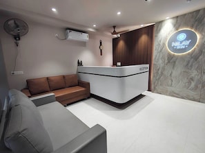 A reception area with a reception desk labeled 'RECEPTION,' a brown leather sofa, a standing fan, and an air conditioning unit.