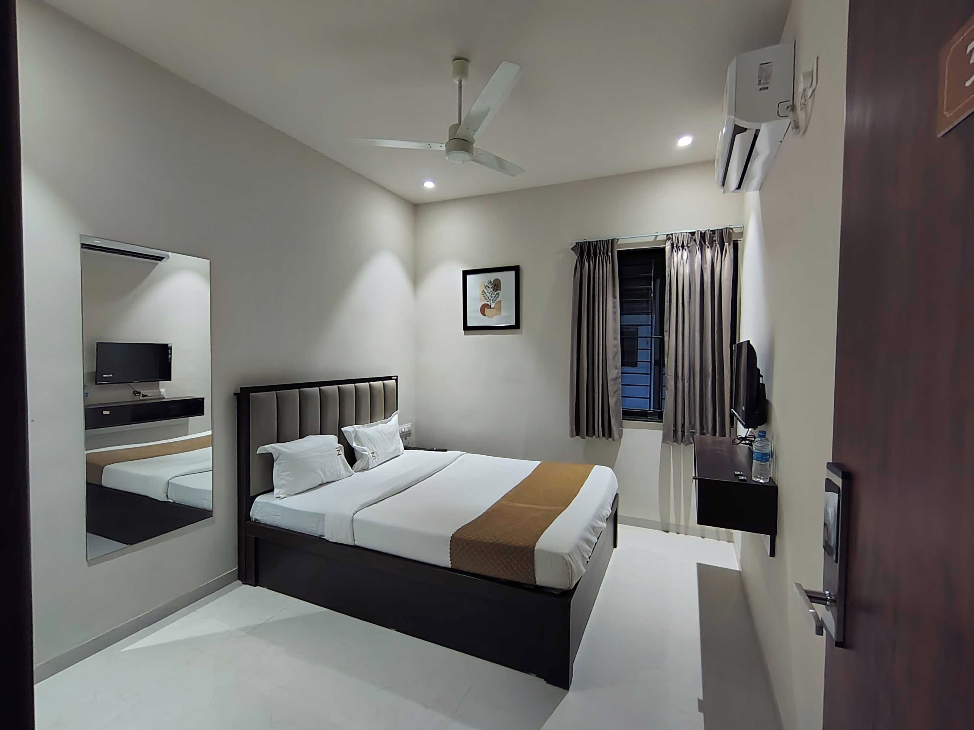 Habitación de hotel con cama, televisor, ventilador de techo, ventana con cortinas y puerta.