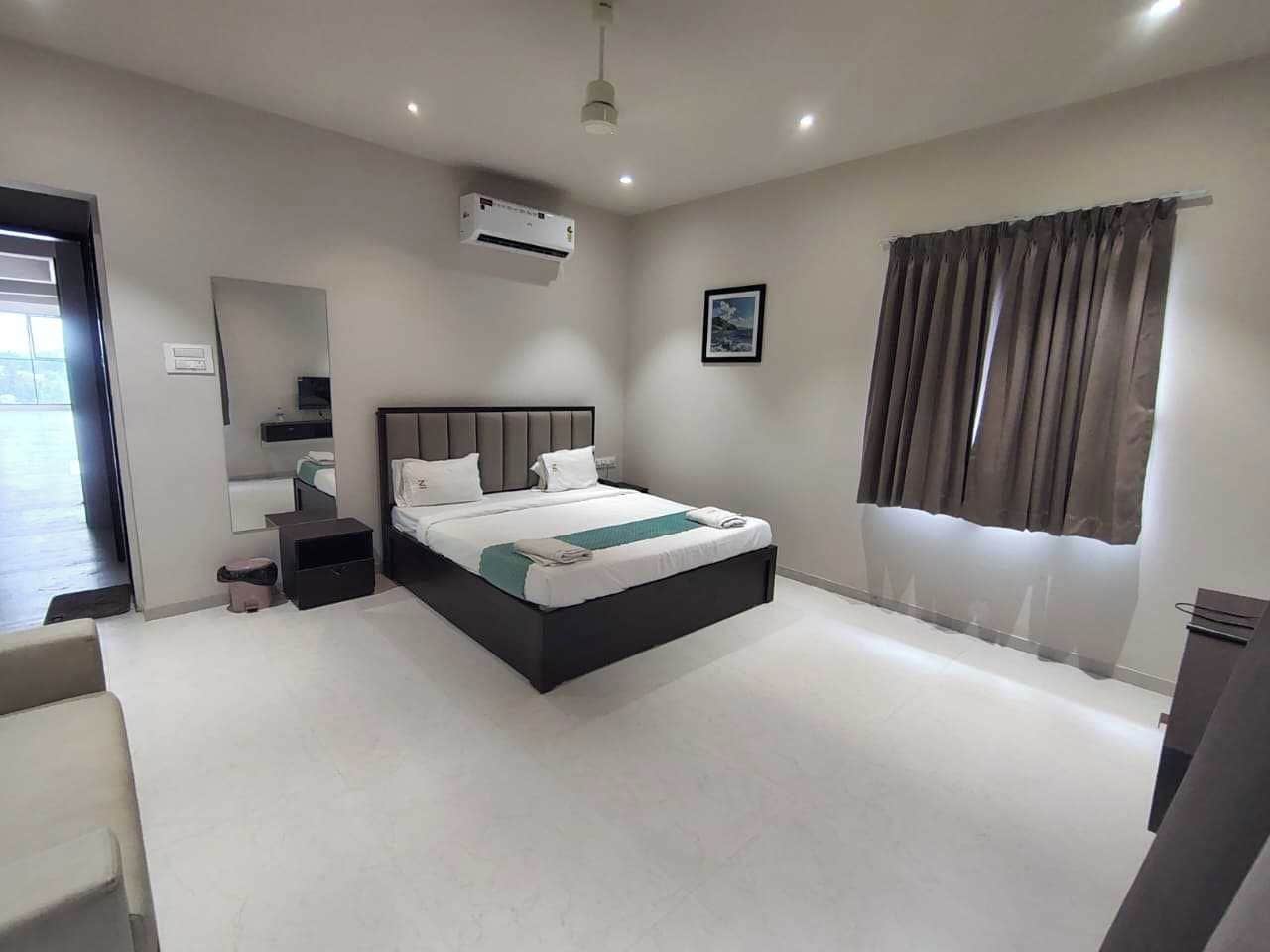A modern hotel room with a large bed, a bedside table, a mirror, an air conditioning unit, and a window with curtains.