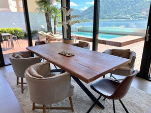 A dining area with a wooden table and chairs, a view of a pool and mountains through large windows. - Luxury Apartment by Interhome (Minusio)