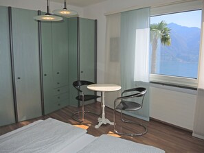 A room with a view of a mountain and a body of water, a small round table, two chairs, and a wardrobe with multiple doors. - Casa Lydia by Interhome (Brione sopra Minusio)