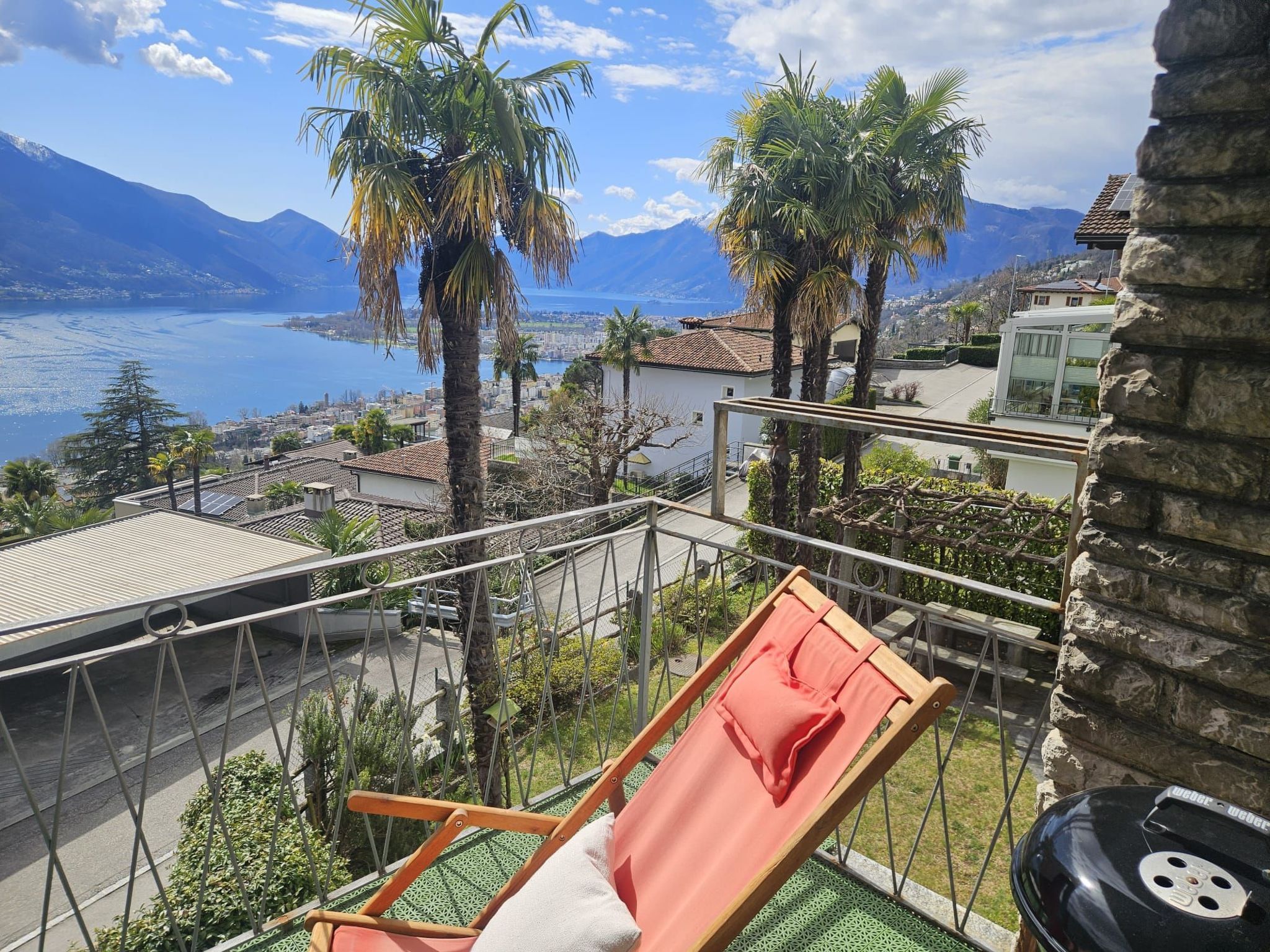 A balcony with a chair and a grill, overlooking a coastal city with mountains in the distance.