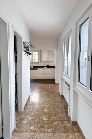 A narrow hallway with a tiled floor leading to a kitchen area with white cabinets and a window. - Villa Genovese by Interhome (Minusio)