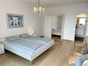 A modern bedroom with a double bed, bedside tables, a chandelier, a mirror, and a view into the bathroom. - Villa Genovese by Interhome (Minusio)