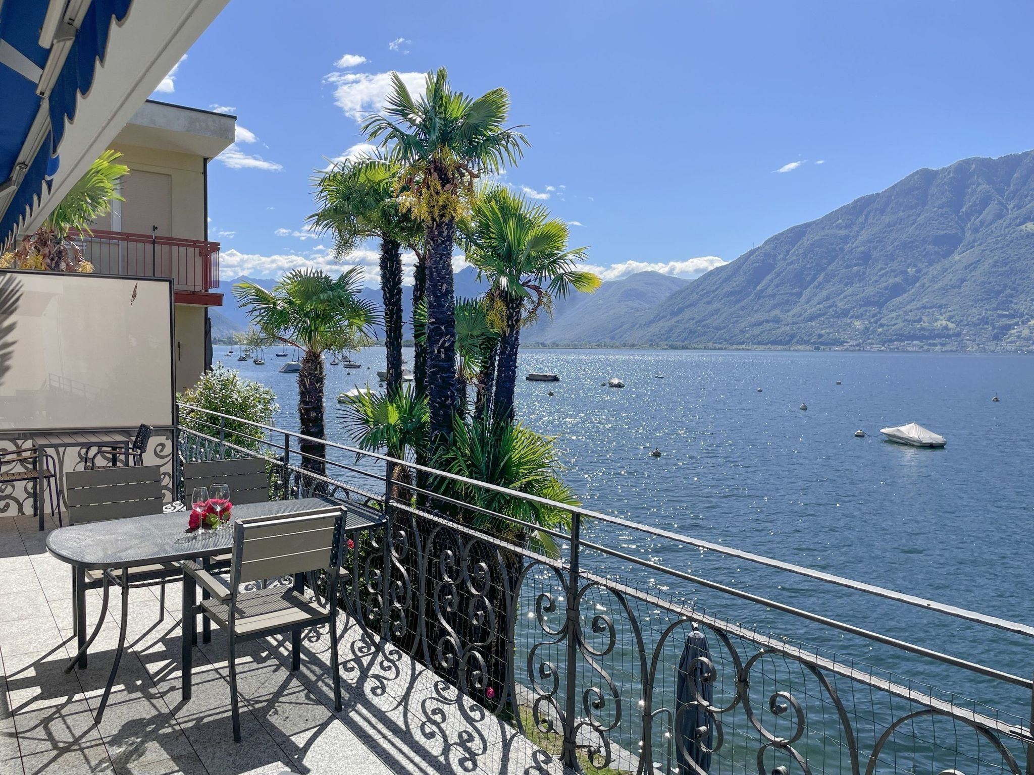 A balcony with a table and chairs, overlooking a lake with boats and mountains in the background.