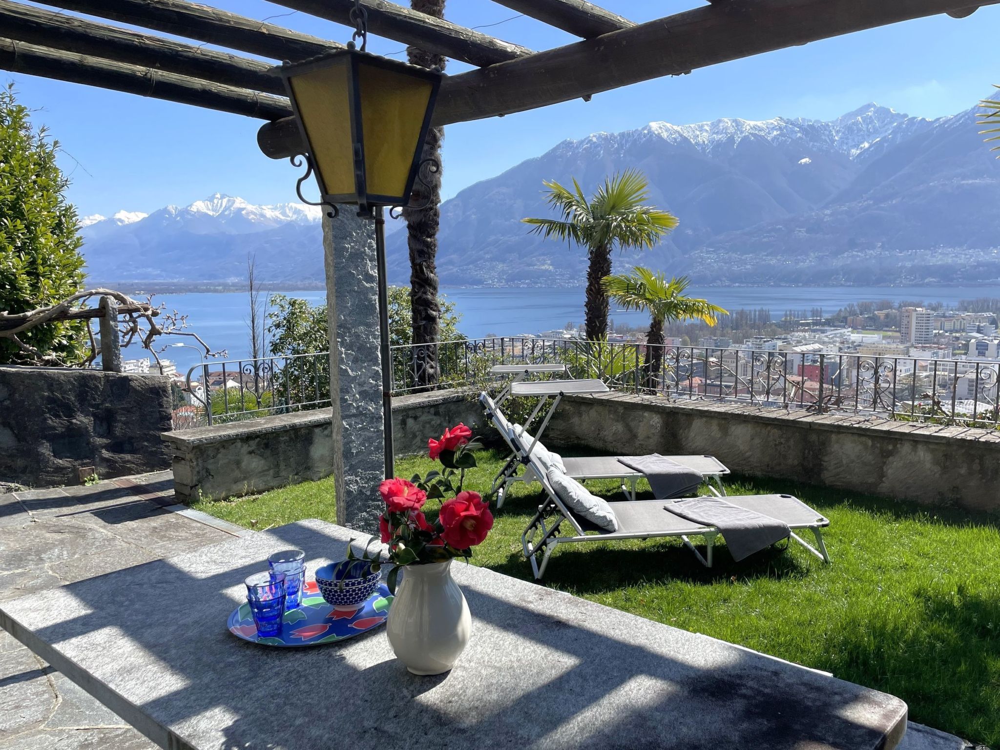 Une terrasse avec vue sur un lac et des montagnes, une table ornée d’un vase de fleurs et des fauteuils de jardin.