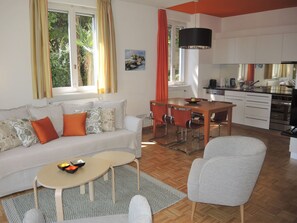 A modern living room with a sofa, dining table, and chairs. There is a kitchen area with appliances and a window with curtains. - Villa Dora Garten by Interhome (Locarno)