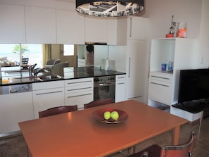 A modern kitchen with white cabinetry, a black countertop, and a dining area with a wooden table and chairs. - Villa Dora Garten by Interhome (Locarno)