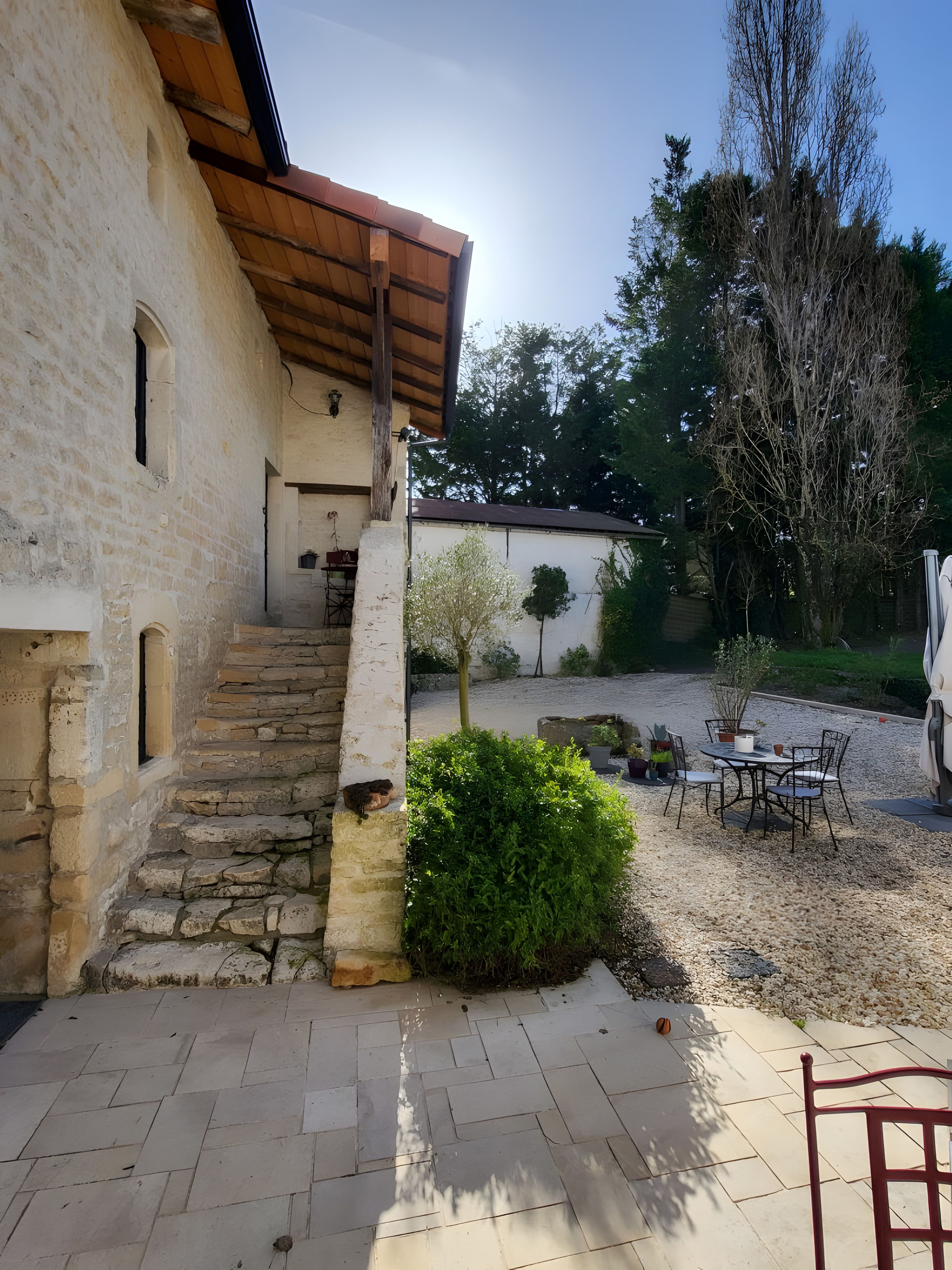 Un escalier en pierre menant à un bâtiment surmonté d’un toit en bois, une terrasse en gravier avec une table et des chaises, et un petit arbre.