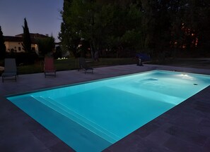 Une piscine rectangulaire, bordée d’une terrasse carrelée, entourée d’arbres et agrémentée de quelques chaises longues.