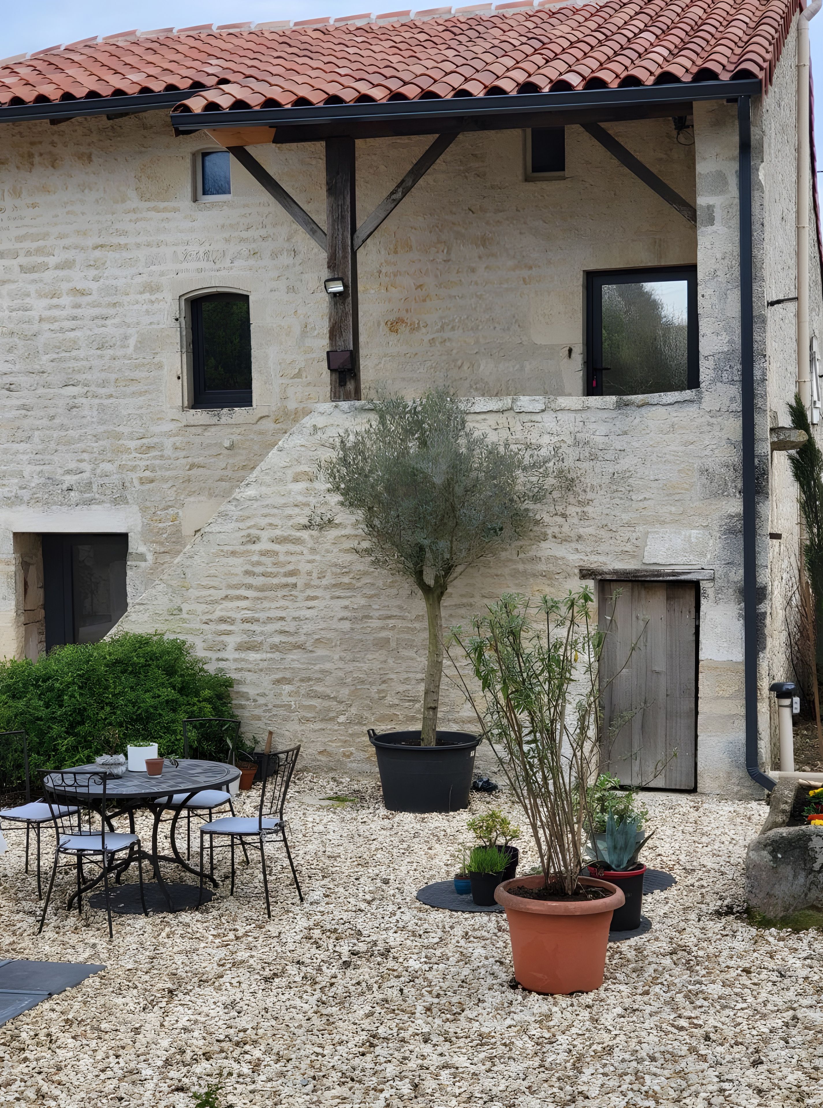 Een stenen gebouw met een pannendak, een klein terras met een tafel en stoelen, en potplanten.