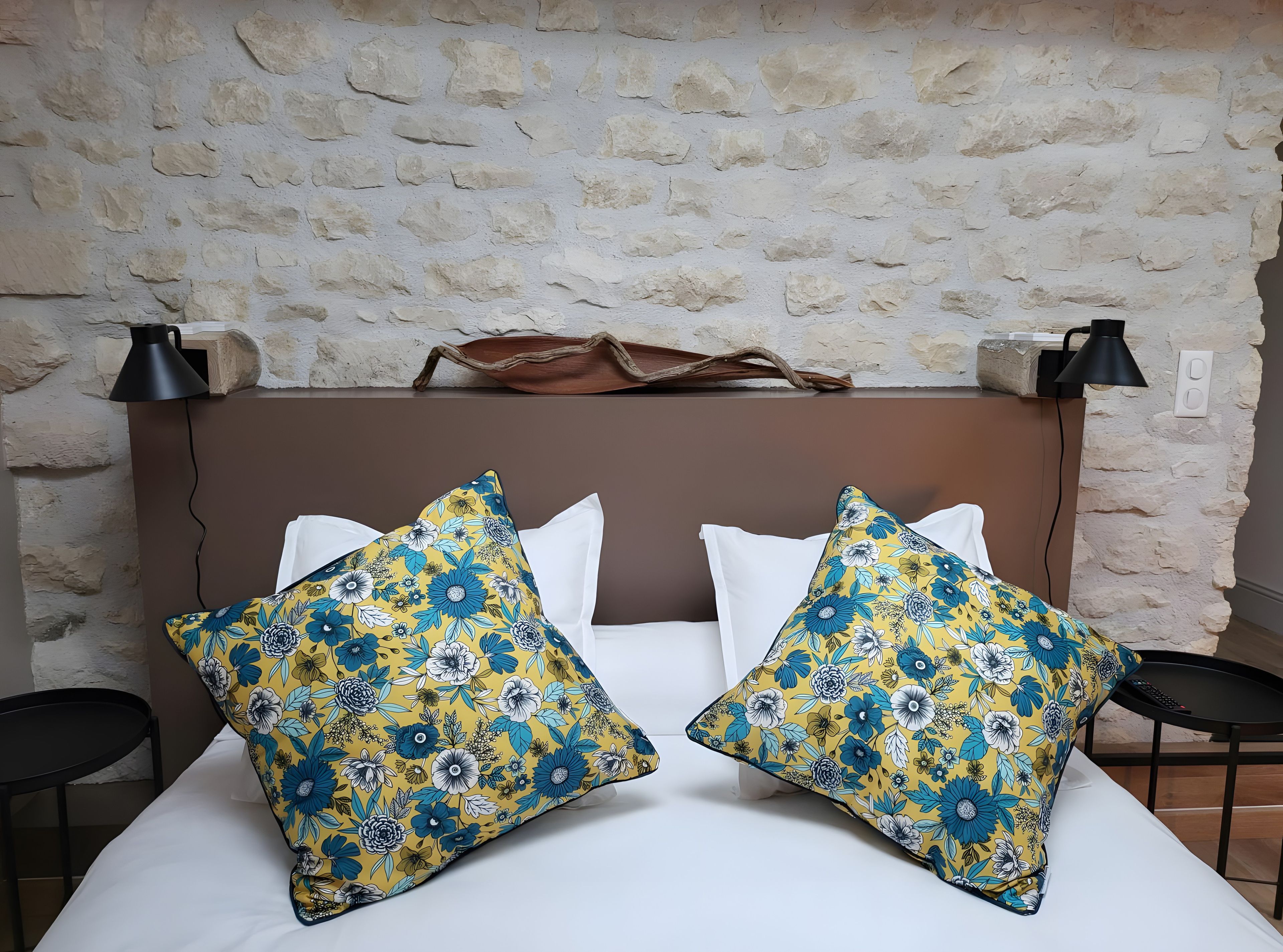 Un lit avec deux coussins décoratifs, un mur en pierre, une lampe et une petite table.
