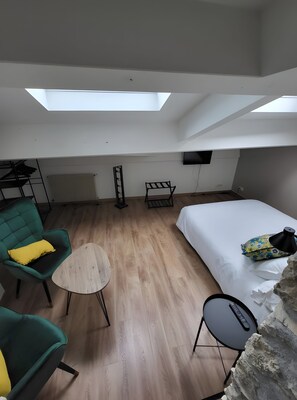 A room with a bed, a green armchair, a small round table, a TV mounted on the wall, and a skylight.
