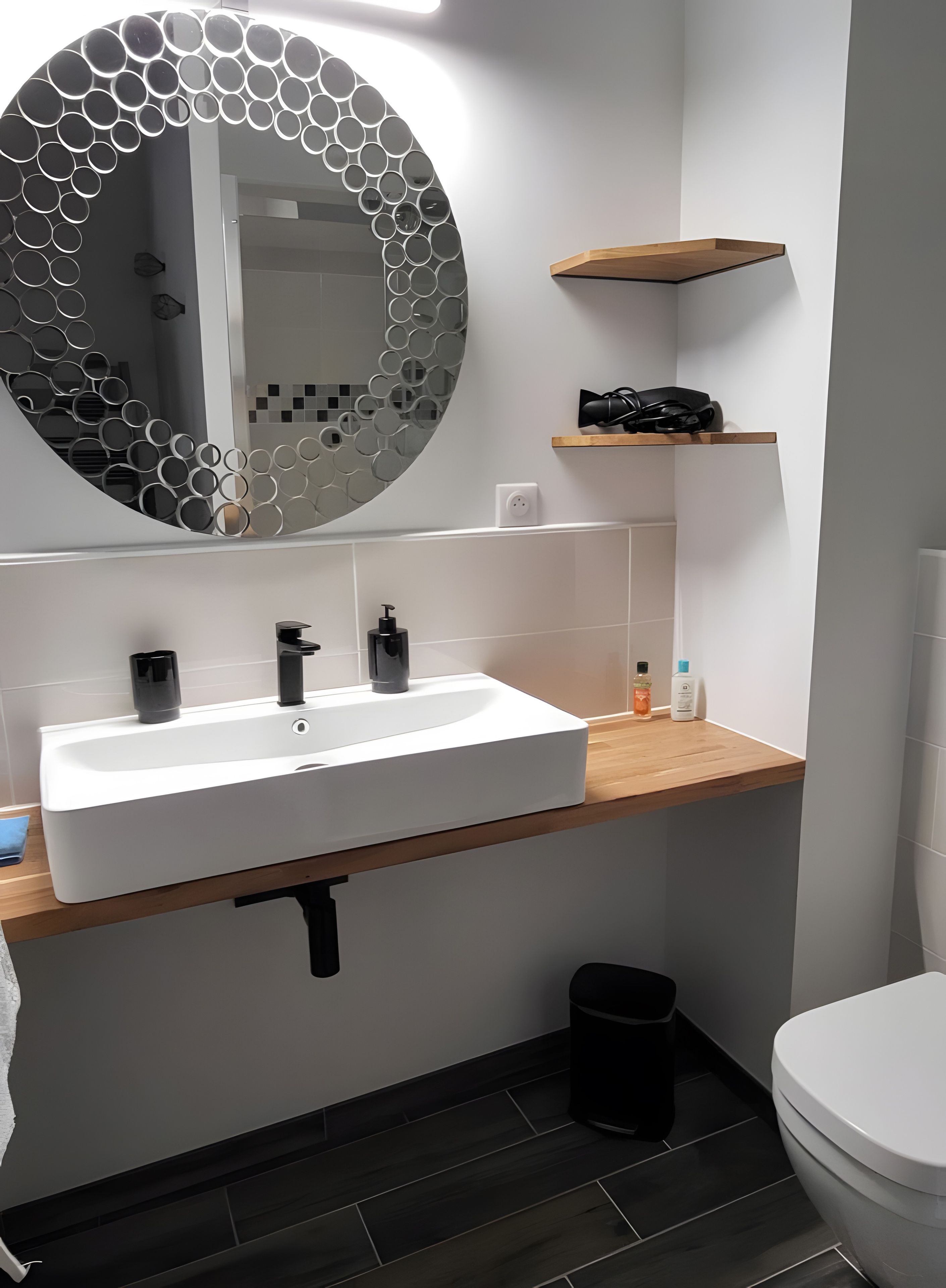 A modern bathroom with a white sink, black faucets, a circular mirror with a honeycomb pattern, a wooden shelf, and a black trash bin.