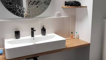 A modern bathroom with a white sink, black faucets, a circular mirror with a honeycomb pattern, a wooden shelf, and a black trash bin.