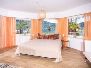 A bedroom with a large bed, two bedside lamps, and a view of a mountain and lake through large windows. - Casa Gioia by Interhome (Gerra)