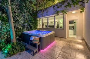Un jacuzzi con un banco y una cesta, rodeado por un muro con ventanas y una puerta que da a una cocina.