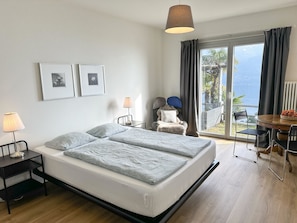 A modern hotel room with a bed, bedside lamps, a small table with chairs, a view of the outdoors, and two framed pictures on the wall. - Casa Rondinella by Interhome (Orselina)