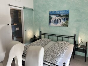 A bedroom with a bed, two bedside tables with lamps, a bathroom with a shower, and a wall-mounted picture of a waterfall. - Casa Makatea by Interhome (Brione sopra Minusio)