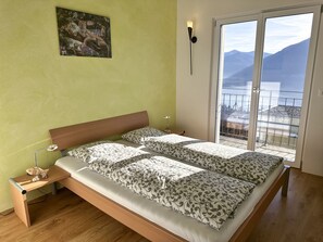 A bedroom with a bed, a nightstand, a lamp, a painting, and a balcony with a view. - Casa Makatea by Interhome (Brione sopra Minusio)