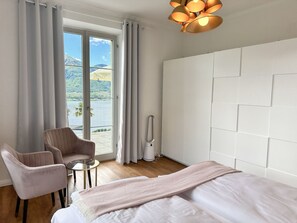 Una camera d'albergo con un letto, una sedia, un tavolino, un termosifone e vista su una montagna e un lago attraverso ampie finestre.