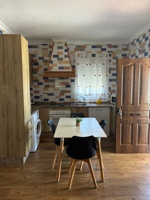 A compact kitchen with a dining table, chairs, a washing machine, and a wooden door. - Casa Luna I        . (El Colorado (Conil de la Frontera))