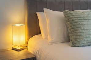 A neatly made bed with white bedding and a green textured pillow, a bedside lamp on a wooden nightstand, and a grey upholstered headboard. - Cosy Apartment in the heart of St Albans. (Free double parking) (St Albans)