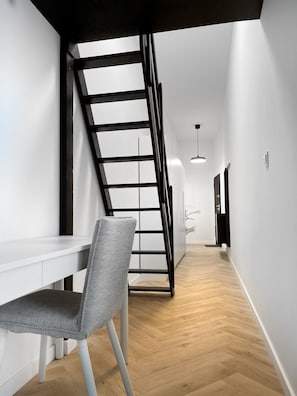 A modern interior with a white desk, a grey chair, a wooden floor, and a black staircase. - Premium Apartment for 3 guest with A/C in Cracow, Dąbrowskiego (Kraków)