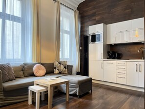 A modern living space with a sofa, ottoman, and a small table. There is a kitchenette with white cabinets and a microwave. - Beautiful Studio for 4 guests, Main Square, Cracow, Marka (Kraków)