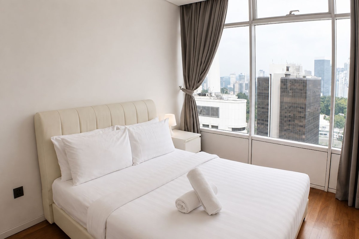 A hotel room with a large bed, a nightstand, a window with curtains, and a view of the cityscape.