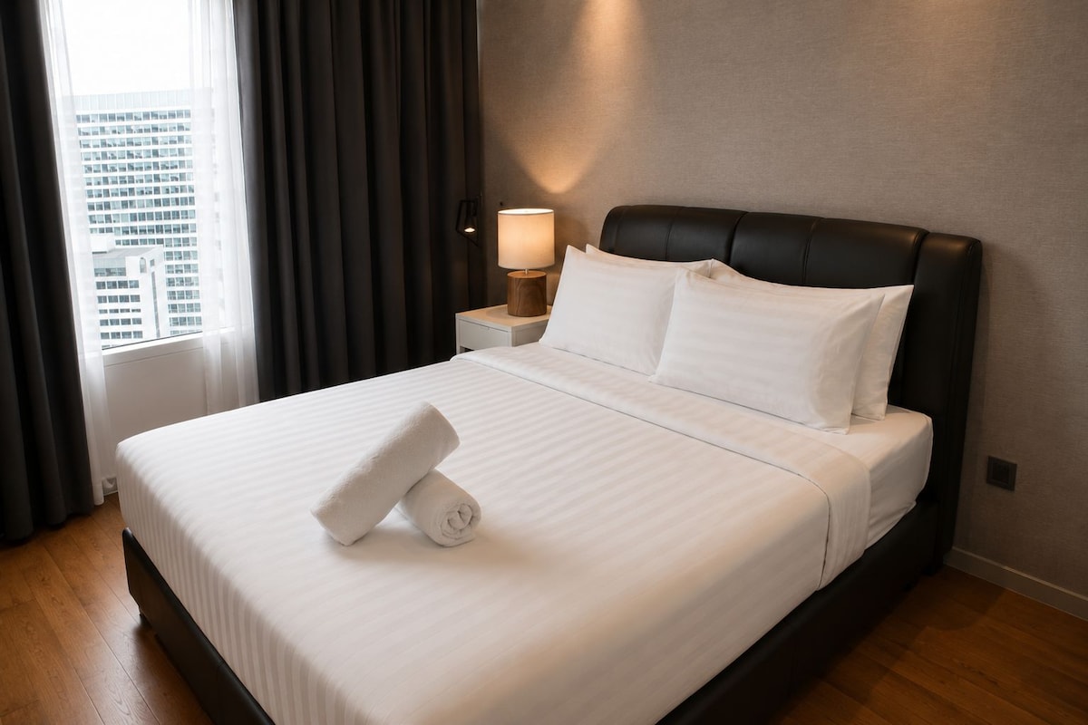 A hotel room with a large bed, a nightstand with a lamp, and a view of buildings through the window.