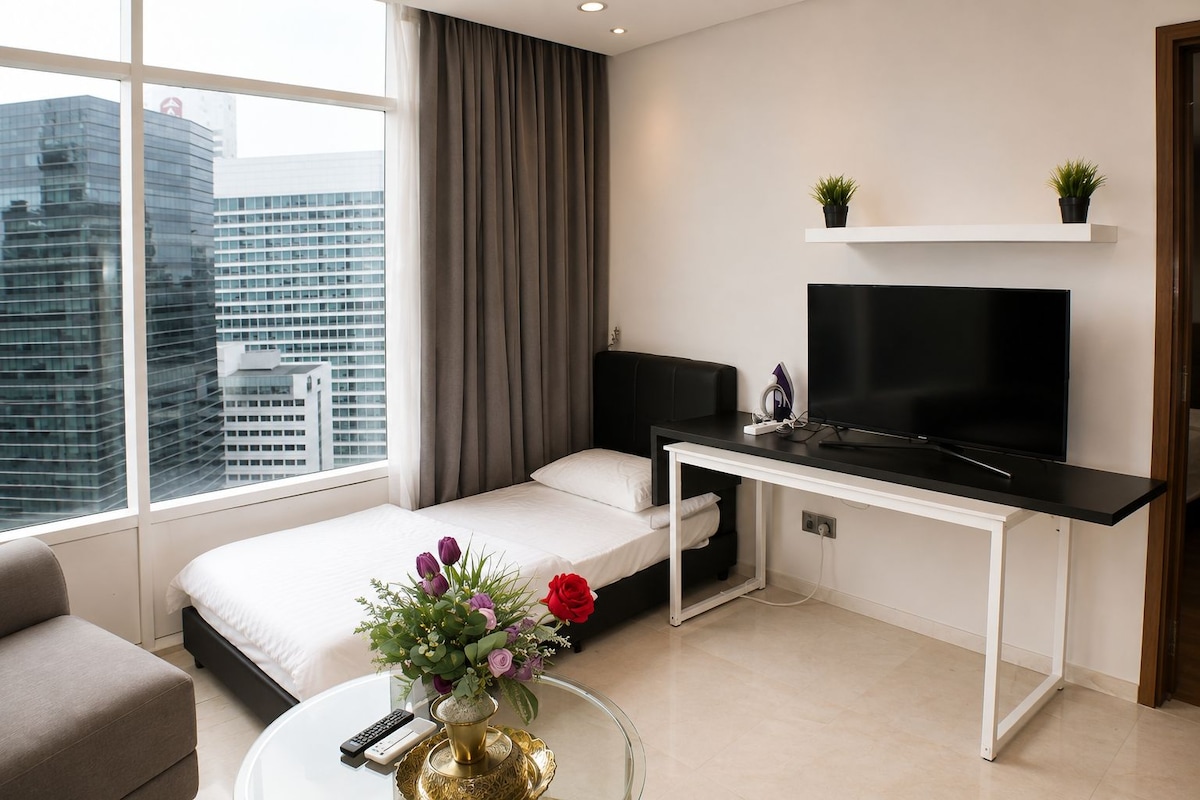 A modern hotel room with a bed, a desk with a flat-screen TV, a small potted plant, and a view of the cityscape.