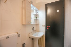 Ein Badezimmer mit einem weißen Waschbecken, einem Spiegel und einer Toilette. Ein Regal mit Toilettenartikeln und eine Tür mit rotem Griff sind ebenfalls vorhanden.