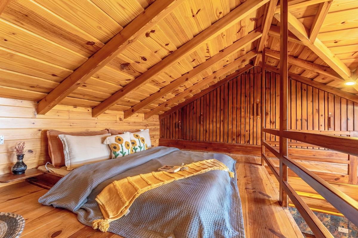 A cozy attic bedroom with a sloped ceiling, wooden beams, a bed with a blue blanket, and a small nightstand with a vase.