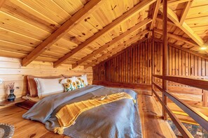 A cozy attic bedroom with a sloped ceiling, wooden beams, a bed with a blue blanket, and a small nightstand with a vase. - Shoonya (Mukteshwar)