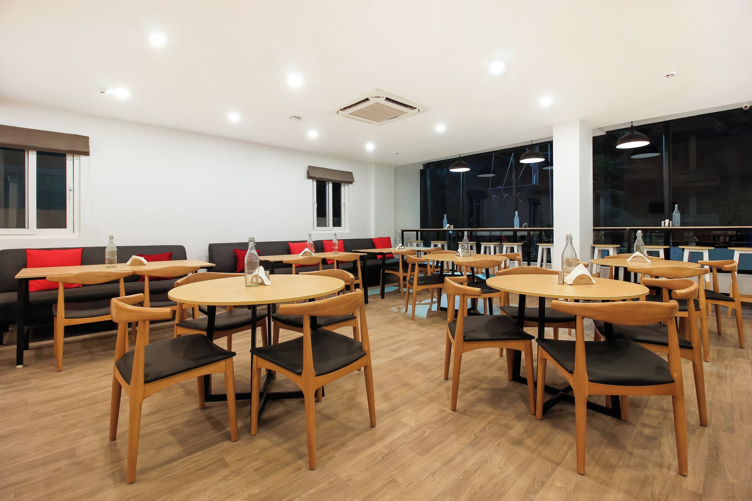 A modern dining area with round tables and chairs, a sofa with red cushions, and large windows with a view.
