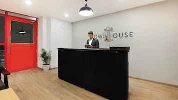 Una mesa de recepción con una persona detrás, una puerta roja, una planta en maceta y una pared con el texto 'owinhouse'.