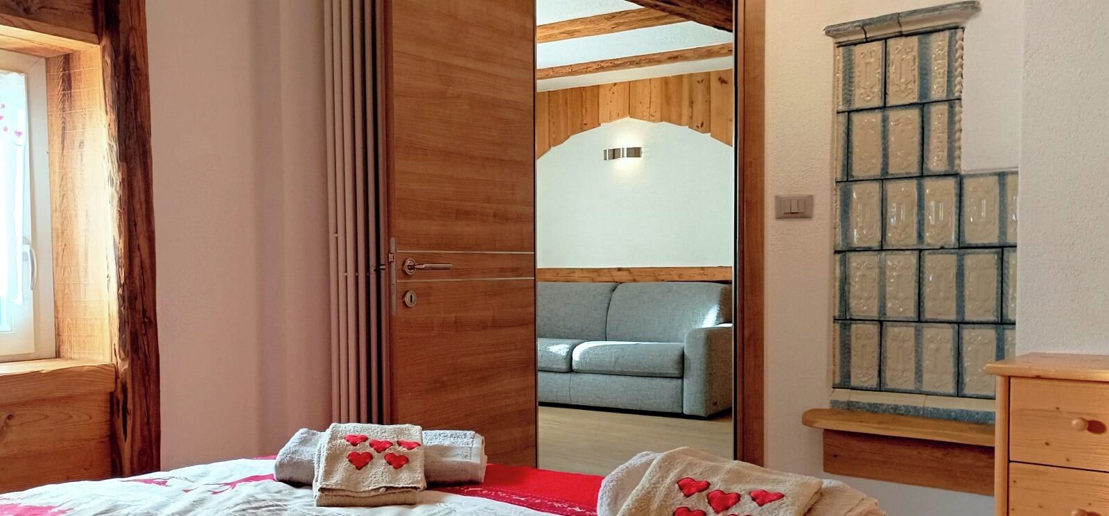 A bedroom with a wooden door, a grey sofa in the adjacent room, and a tiled wall.