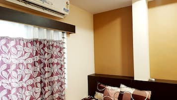 Una habitación de hotel con cama, ventana con cortinas, unidad de aire acondicionado y estante montado en la pared.
