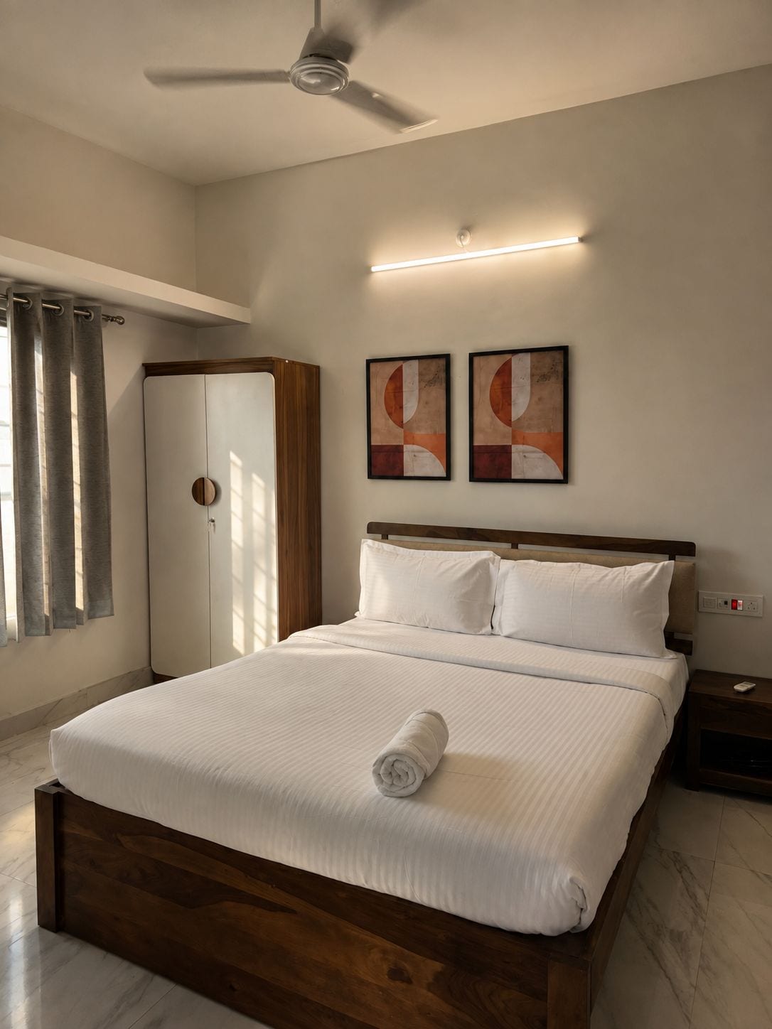 Habitación de hotel con cama, ventilador de techo, mesita de noche, armario y dos cuadros abstractos en la pared.